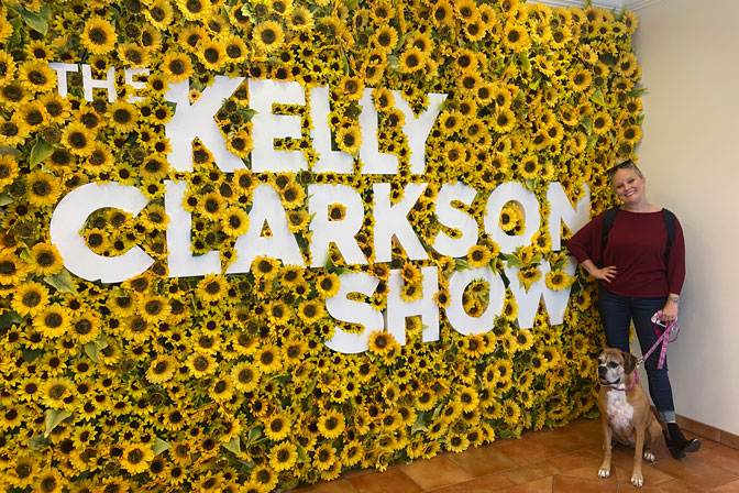 Camille Cox with her dog pose in front of The Kelly Clarkson Show sign