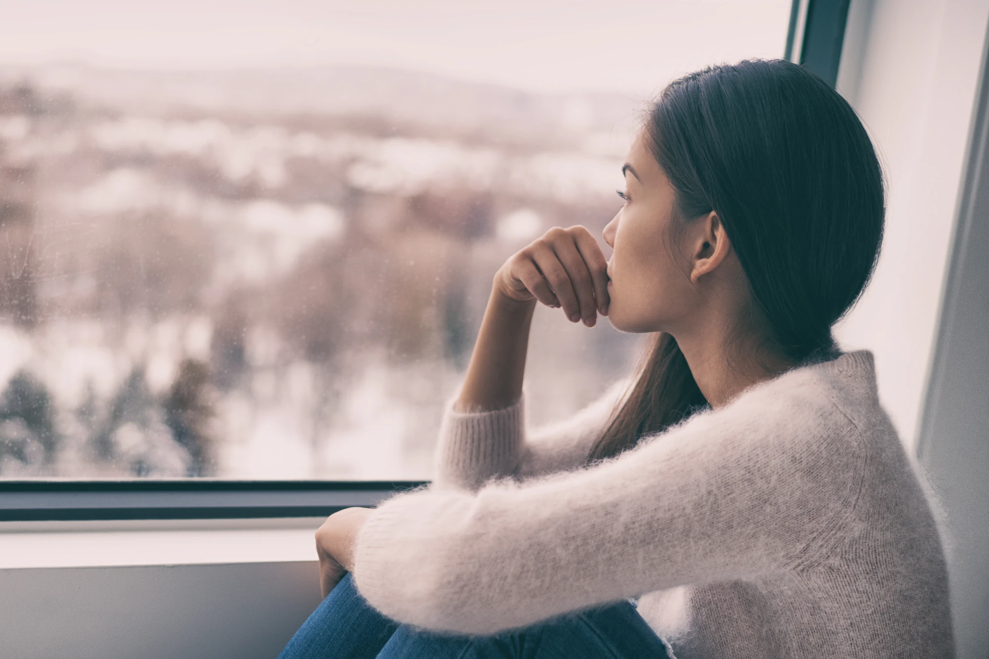 A woman sitting by a window at home, looking outside with a thoughtful expression