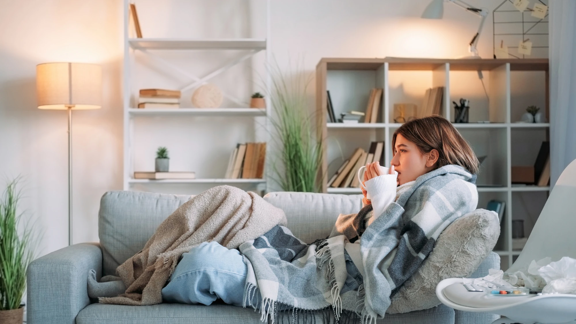 A young woman sitting at home wrapped in a soft blanket, looking down with a quiet expression