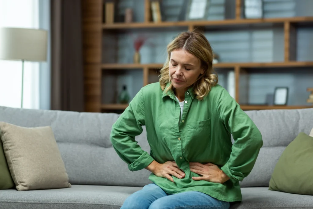 A woman sitting on a couch at home with her hand on her stomach, looking uncomfortable